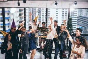 Business team celebrating at an office New Year party with confetti, party hats, and employees cheering together.